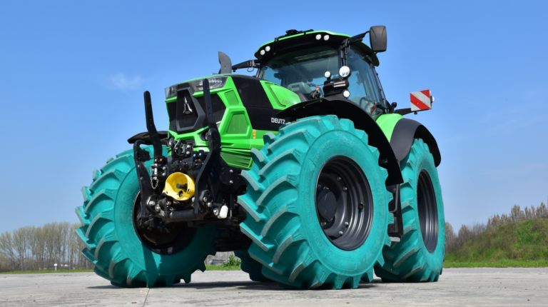 A tractor fitted with green GRI Tires exstands parked on a tarmac road with green bushes behind it and a blue sky above
